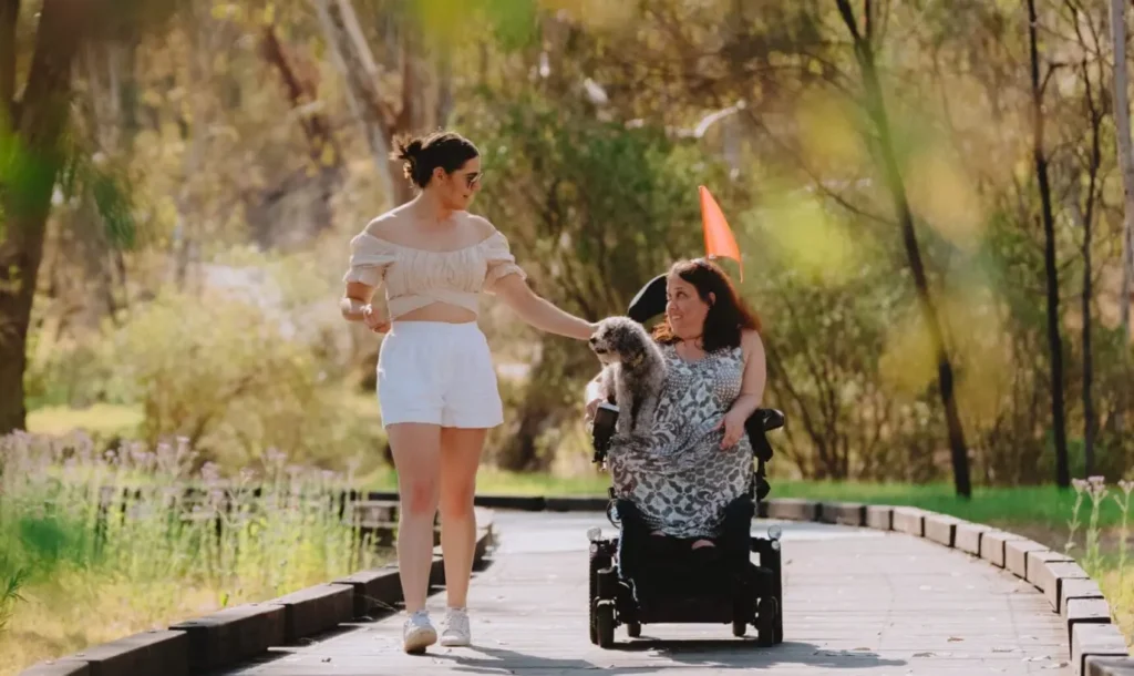 A woman in a wheelchair smiles while holding a small dog, alongside a friend walking beside her on a leafy, accessible trail in Albury Wodonga.