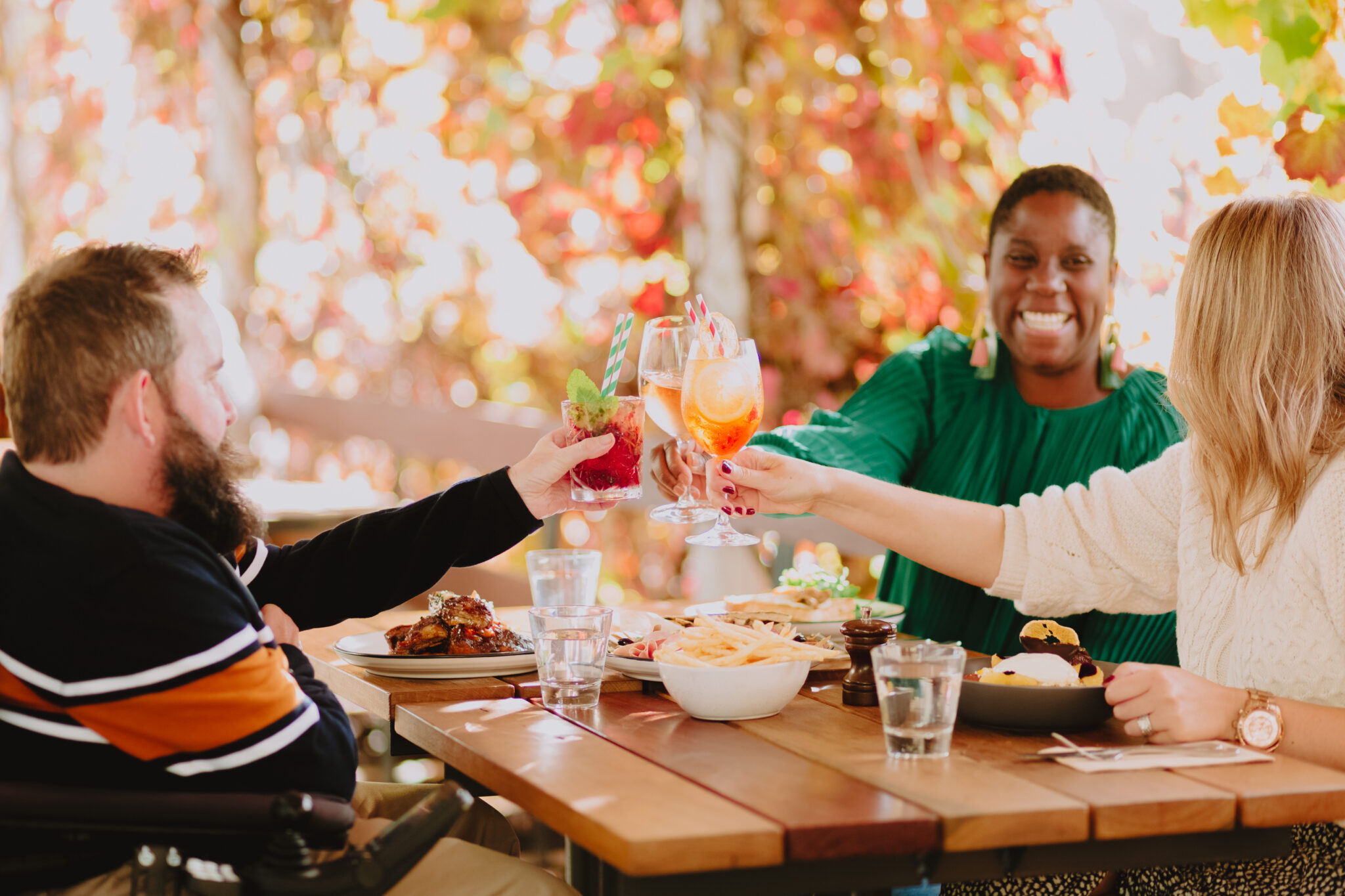 Man in electric wheelchair sits opposite two women at cafe table under autumn-coloured grapevines. They hold their drinks up to cheers.