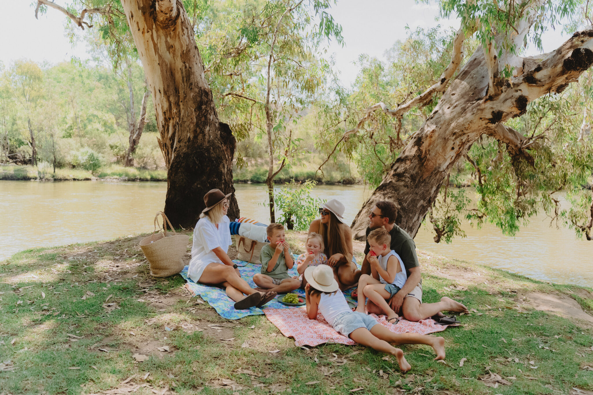 A family enjoying a riverside picnic under the shade of eucalyptus trees along the Murray River in Albury Wodonga, with children eating fruit and adults relaxing on a blanket.