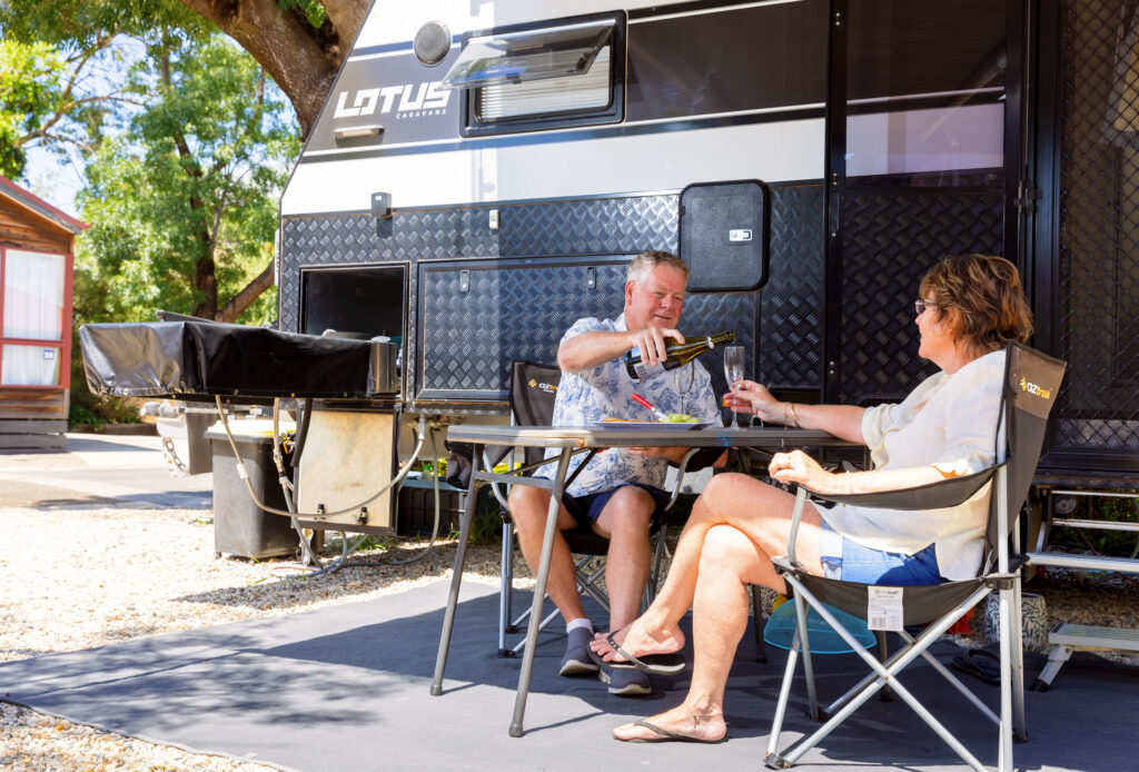 Older couple sit at fold out picnic table in front of caravan. The man pours a glass of wine