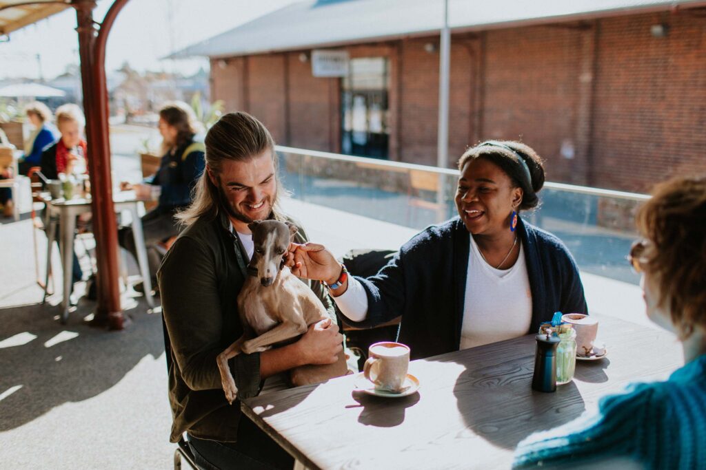 A smiling man holds a dog on his lap while sitting at a cafe table with two women drinking coffee