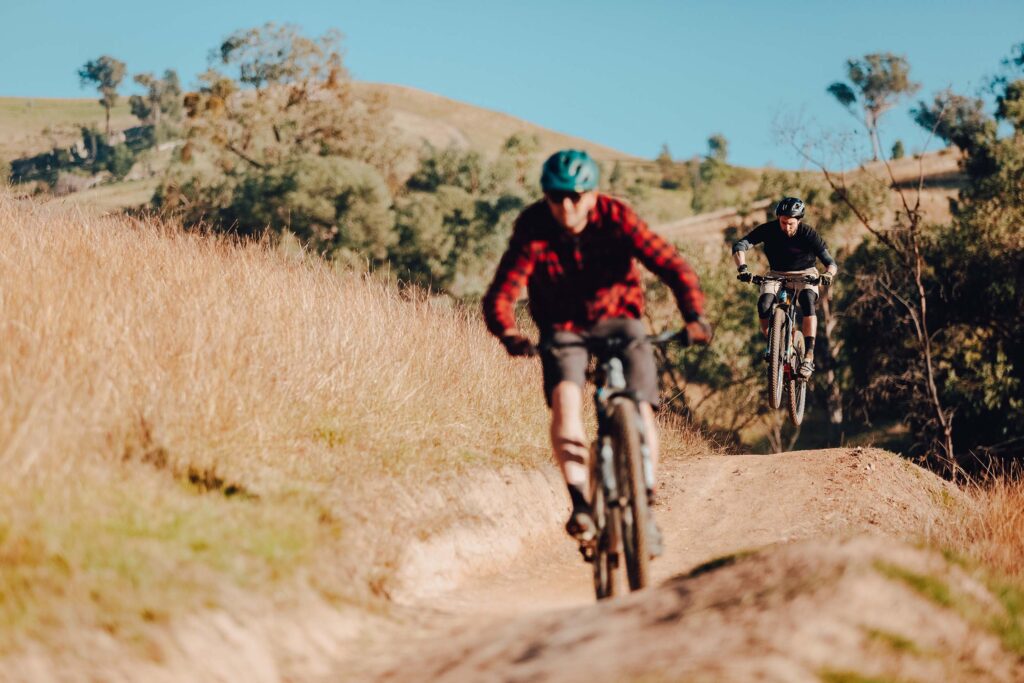 A cyclist in a red plaid shirt and green helmet rides on a dirt track at McFarlanes Hill. Another rider in black becomes airborne as he comes over a rise.