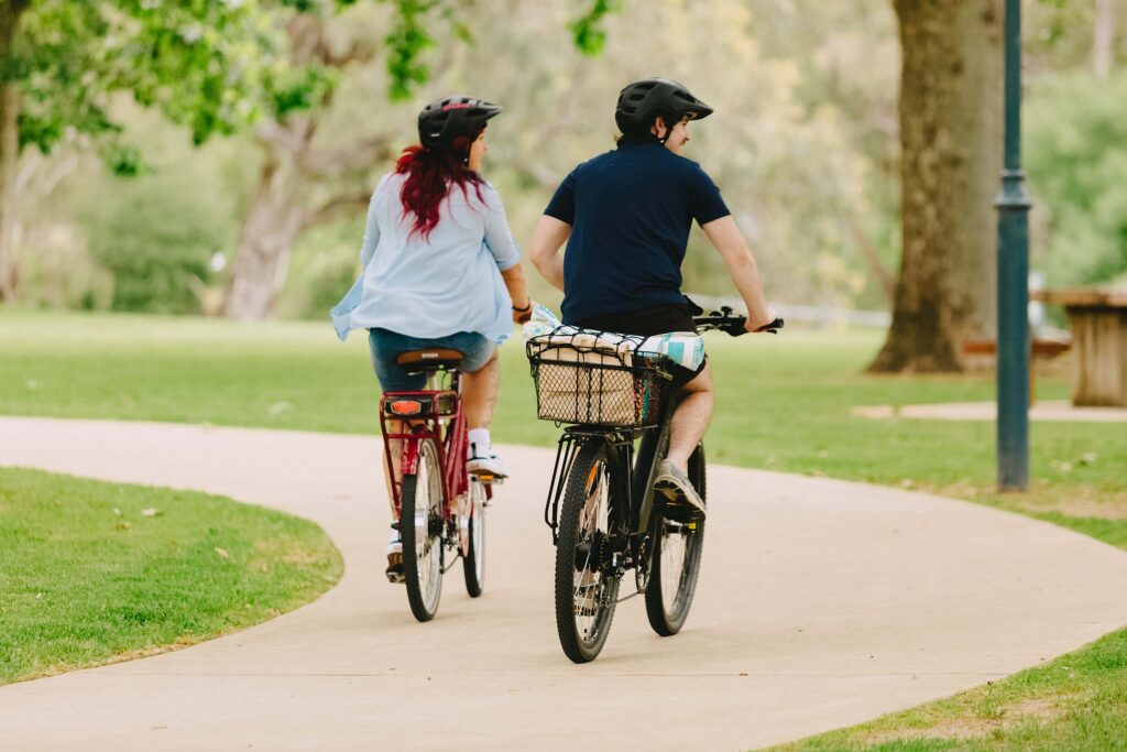 A young man and woman ride bikes along footpath through a grassy park
