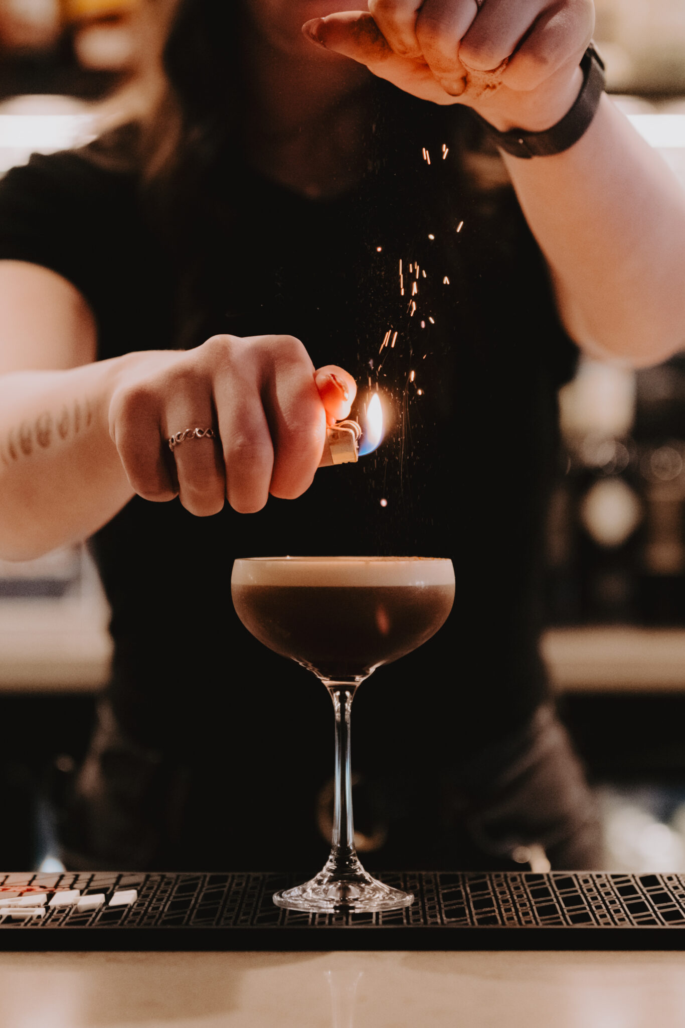 Woman in black t-shirt holds lighter over espresso martini on bar mat, sprinkling garnish onto drink and creating sparks