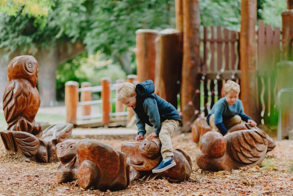 Two young boys play on large wooden animal carvings in a nature-themed playground surrounded by trees and autumn leaves.