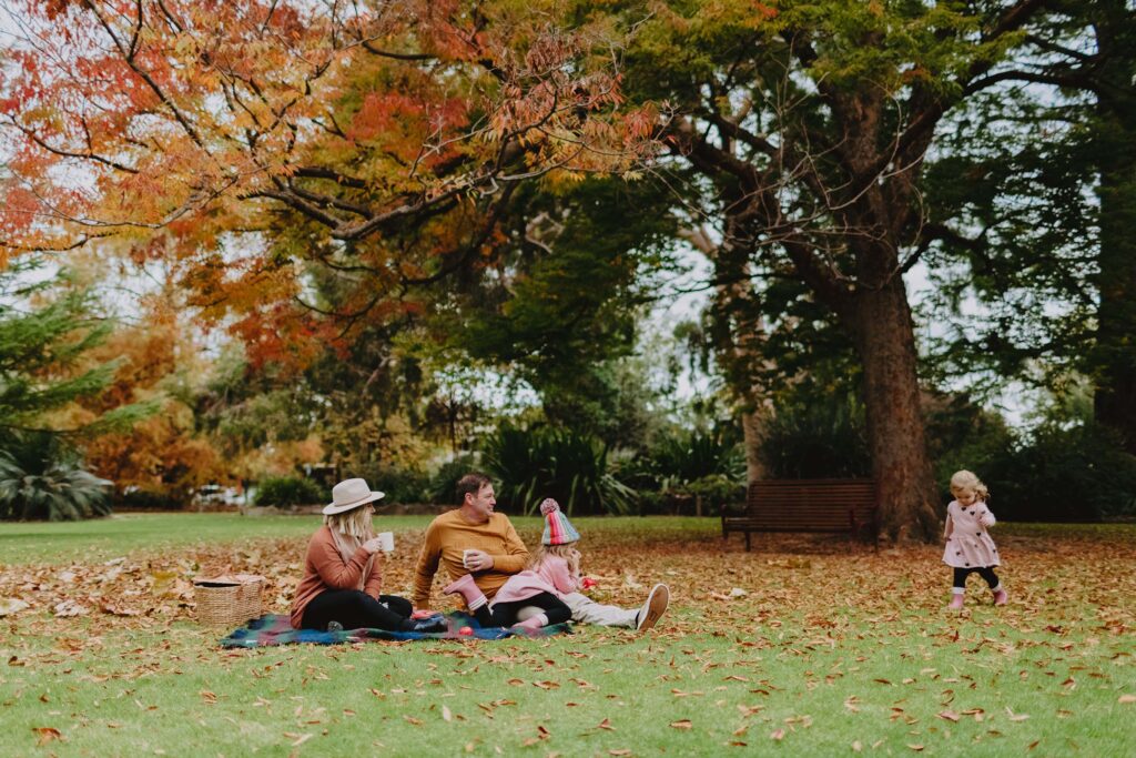 A family sit on a picnic rug underneath autumn trees at the Albury Botanic Gardens. A little girl in a pink dress runs through the fallen leaves.