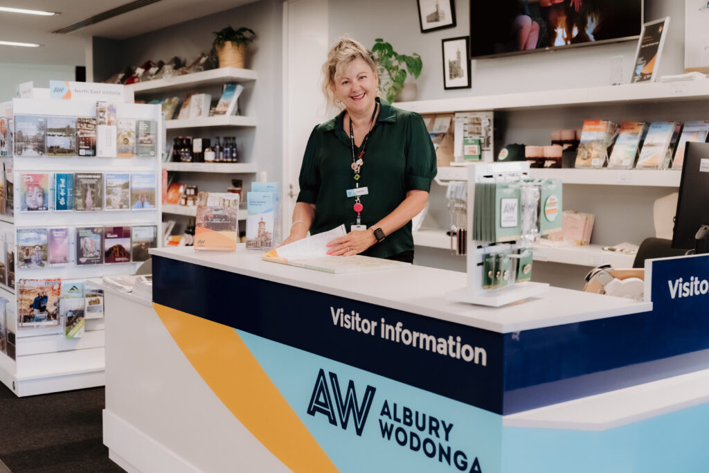 Blonde woman with a welcoming smile wears a green blouse and a lanyard. She is standing behind a desk that reads 'visitor information' with the 'AW Albury Wodonga' logo. There are souvenirs on the counter, and a brochure stand in the background.