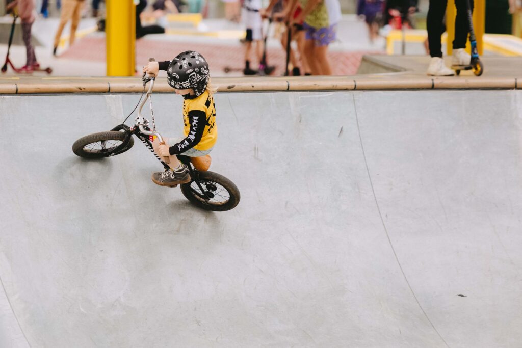 A young child wearing a helmet rides a small bike around the curve of a skate park bowl, with other kids on scooters in the background.
