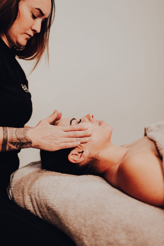 A woman in a black shirt placing her hands on the face of another woman laying down on a massage bed.