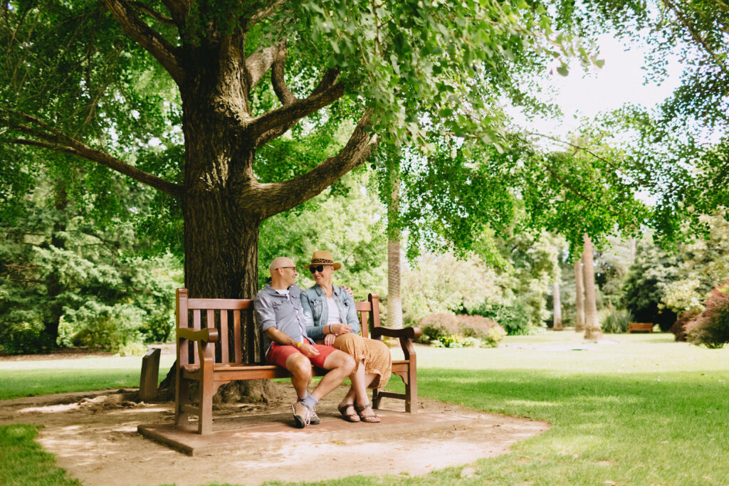 A couple sits closely together on a wooden bench under a large, leafy tree in a sunny park. The man wears a light gray shirt and red shorts, while the woman is dressed in a denim jacket, a white shirt, a patterned skirt, and a hat. Both are smiling warmly at each other, enjoying the peaceful green surroundings with lush grass and trees in the background.