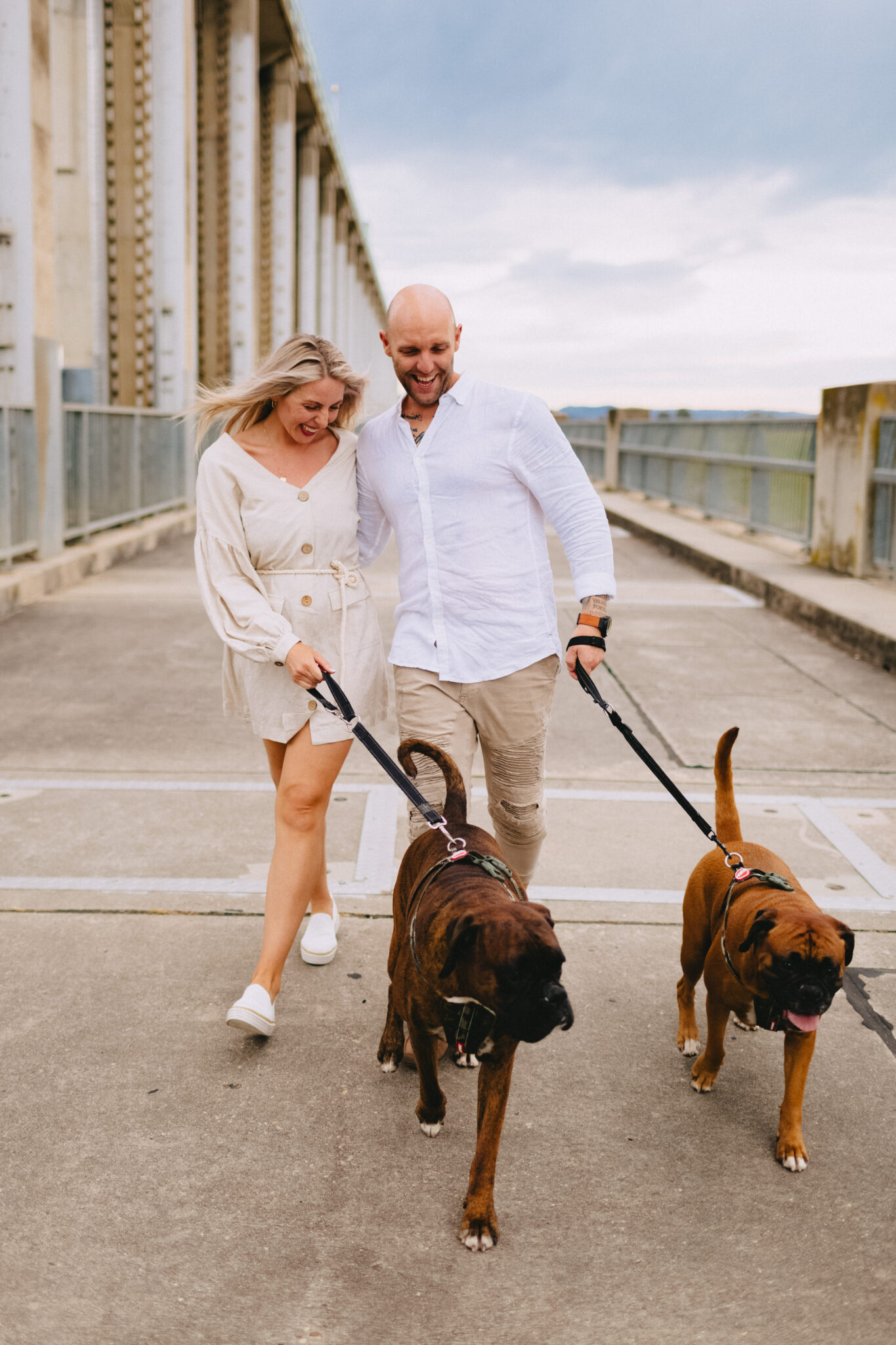 A man and a woman wearing white are walking on a concrete path with two brown coloured dogs on black leads