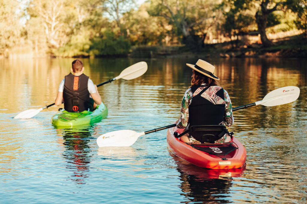 A woman in a red canoe and a man in a green canoe are paddling in water. The woman is wearing a hat and both the man and the woman are wearing life jackets