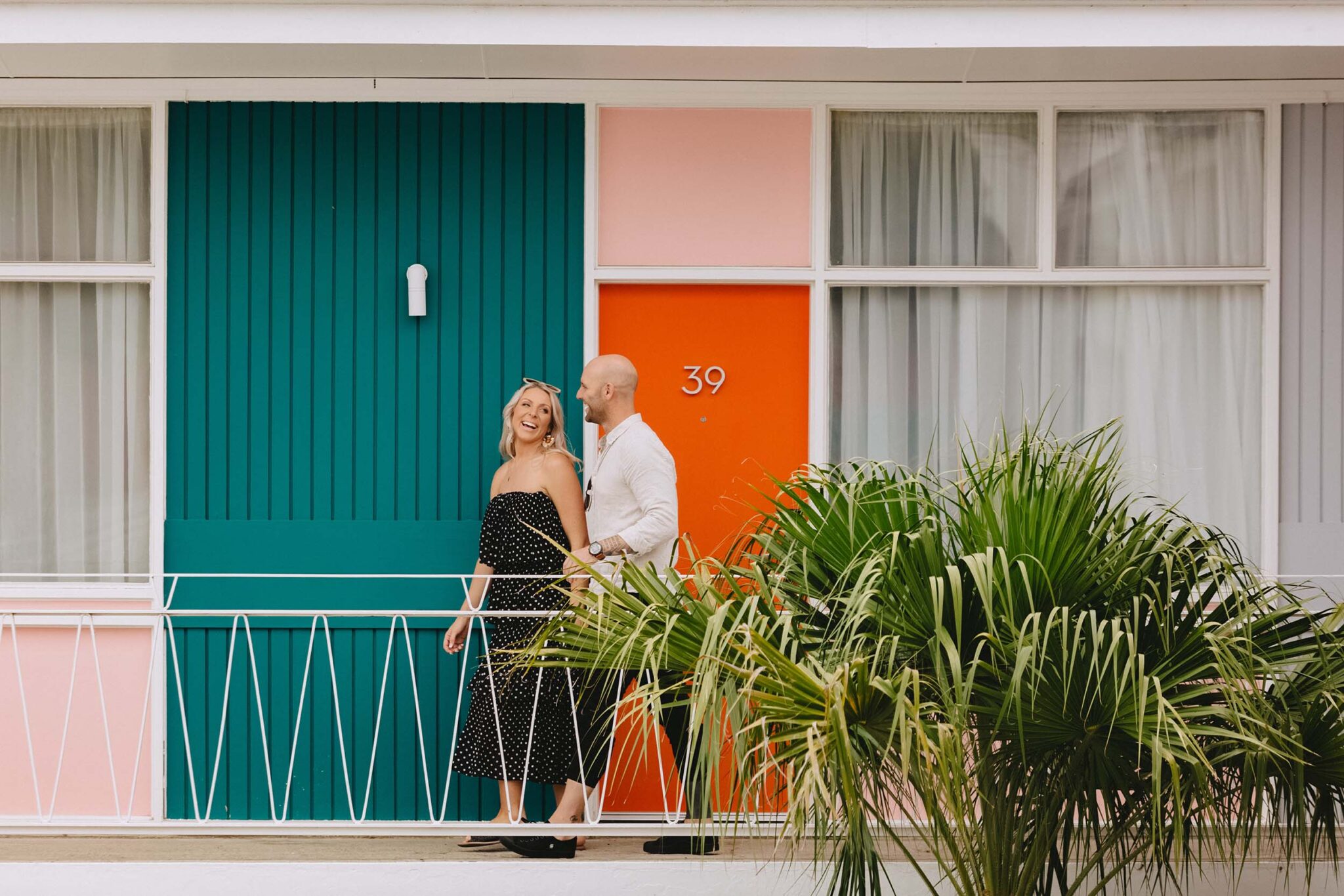 A blonde woman in a black polka dot dress and a man in a white shirt walk along a raised pathway in front of a colourful hotel with a green wall and orange door, and palm fronds in the foreground