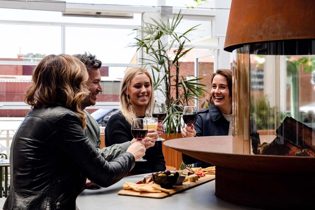 A group of friends drinking wine and beer hold their glasses us to cheers over a share platter at a table with a wood-fire heater in the centre. There is an indoor plant in the background, and the sky outside is grey.