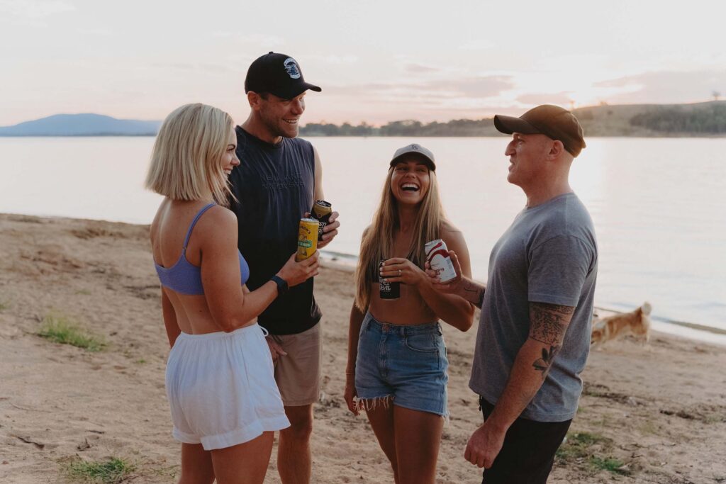 Four friends laughing and holding drinks on a sandy lakeside beach at sunset, with calm water and soft evening light in the background.