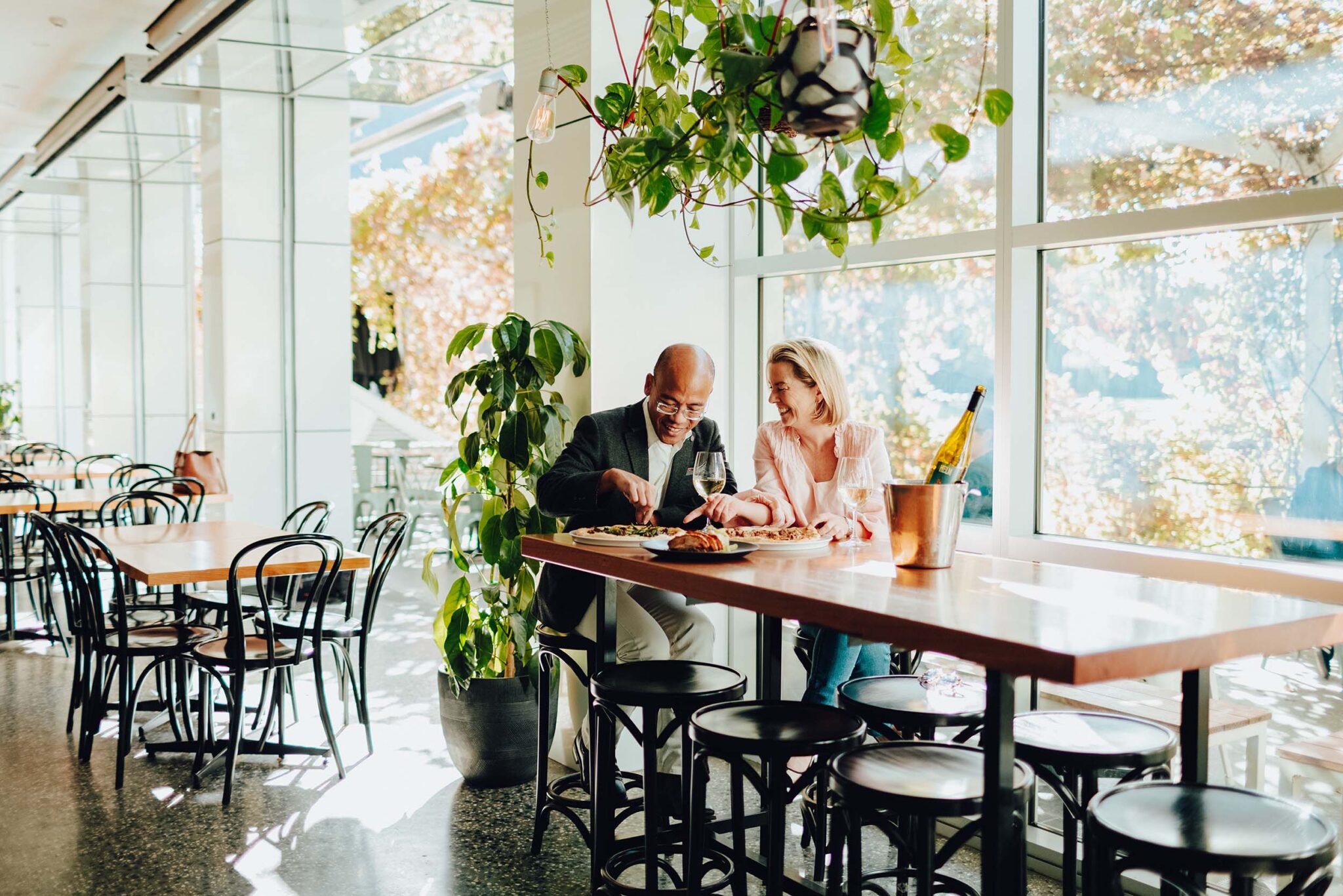 A man in a grey blazer and a woman in a pink blouse dine in a light and spacious cafe with indoor greenery