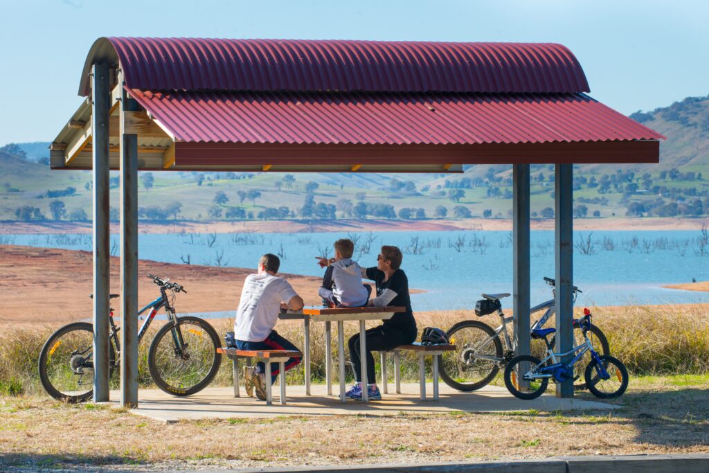 Bikes lean against the poles of a picnic shelter with a red corrugated iron roof. A family sits at the picnic table looking out over Lake Hume, where tree branches can be seen sticking out above the water level.