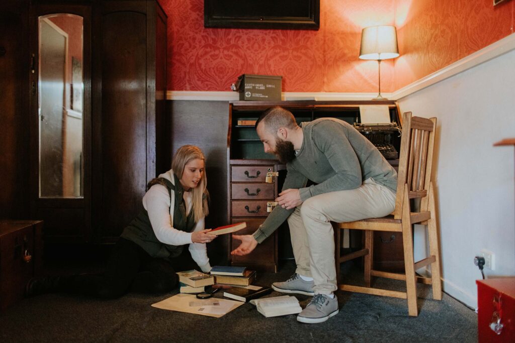 A woman sits on the floor of an escape room next to a pile of books. A man sitting on a wooden chair leans down to look at a book she is handing him