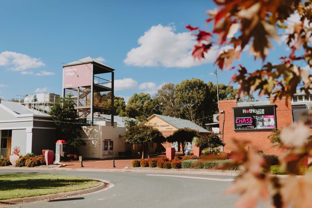 Exterior view of Gateway Village in Albury Wodonga on a sunny day. Featuring the HotHouse Theatre building, a red brick building with a sign promoting a show, trees in the background, and autumn leaves in the foreground.