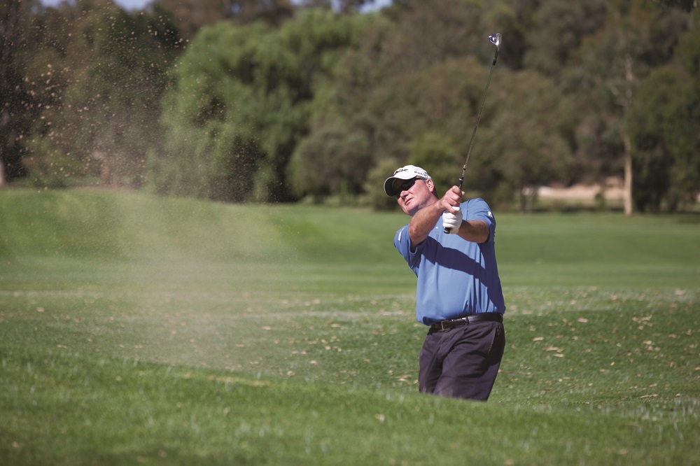 Man in blue polo shirt and white cap on a golfing green with trees in the background holds golf club high over his shoulder at the tops of his swing. His eyes are on the golf ball and a cloud of dirt hangs in the air.