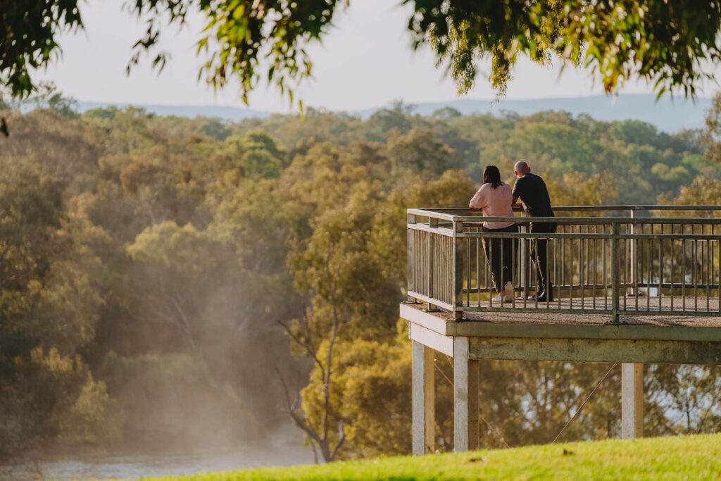 A man and a woman lean on the railing of a platform overlooking the Murray River and a canopy of eucalypt trees