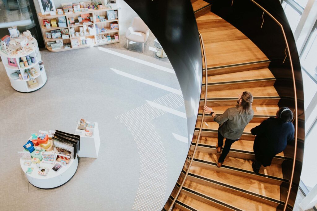 A man and woman walk up the wide spiral staircase of a public library that has white display units in the giftshop on the ground floor