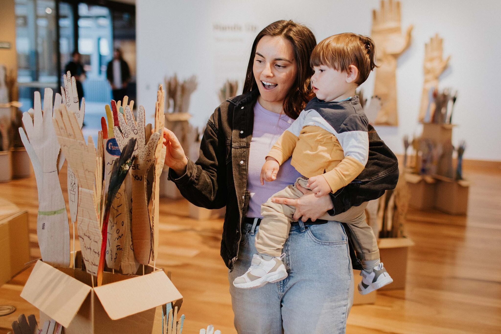 A woman holding a young child explores a hands-on art installation made of cardboard at Hyphen – Wodonga Library Gallery, encouraging creative play and family-friendly discovery.
