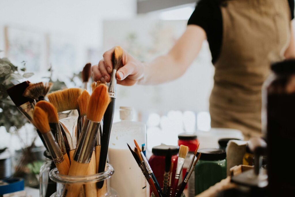 Close-up of an artist's hand reaching for a paintbrush among a collection of brushes in jars, with various paints and art supplies on a table.