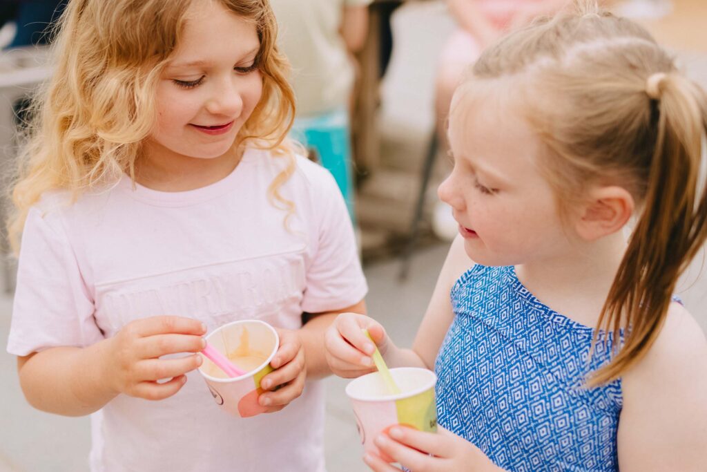 Two young girls enjoy cups of ice cream while chatting outdoors on a warm day, one in a pink t-shirt and the other in a blue patterned dress.