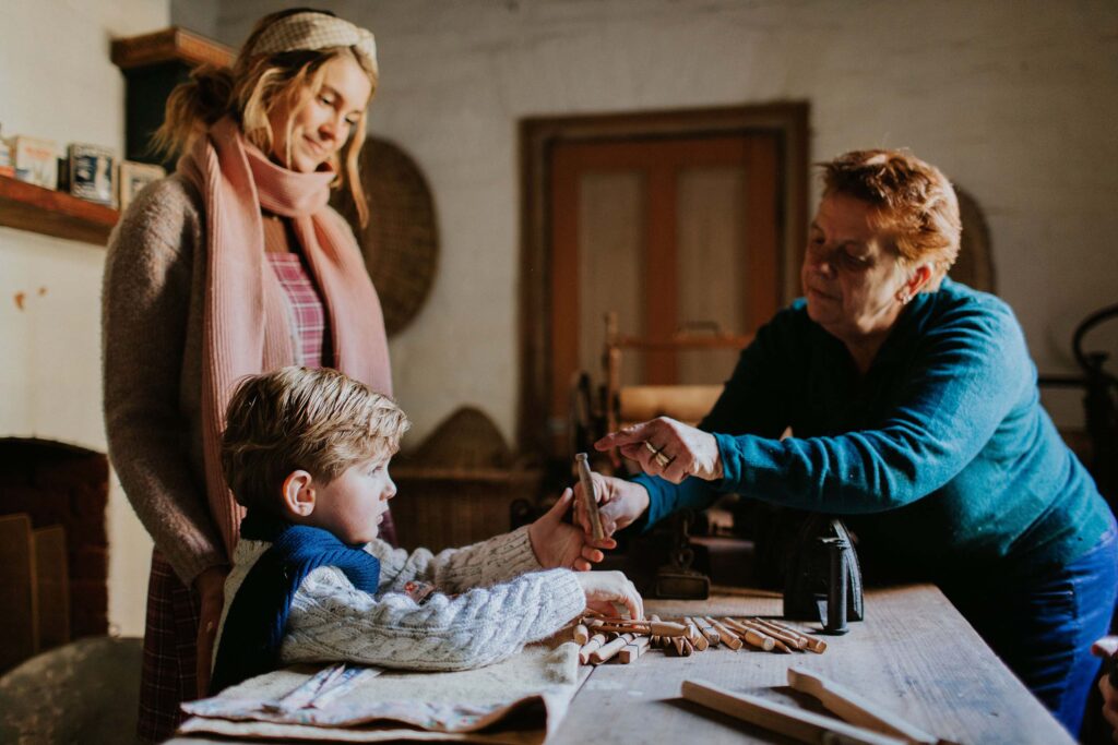 A young boy learns to use old-fashioned wooden pegs at a historical museum, guided by an older woman, while his mother watches over him.