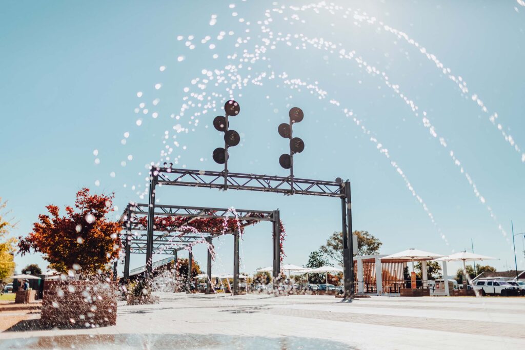 Streams of water from a fountain arc in front of the railway gantry at Junction Place, Wodonga, set against a clear blue sky. The grapevine growing over the gantry has turned a vivid Autumn-red