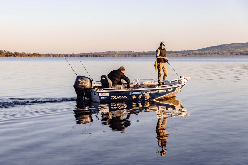 Two people fishing from a motorboat on a calm lake during golden hour, with reflections on the water and distant tree-lined shore.