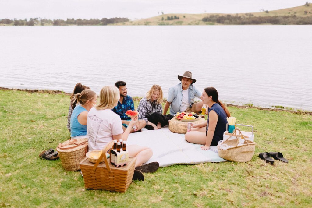 A group of friends sit around the edge of a picnic blanket on the grassy foreshore of Lake Hume. They have wicker baskets and a fruit platter
