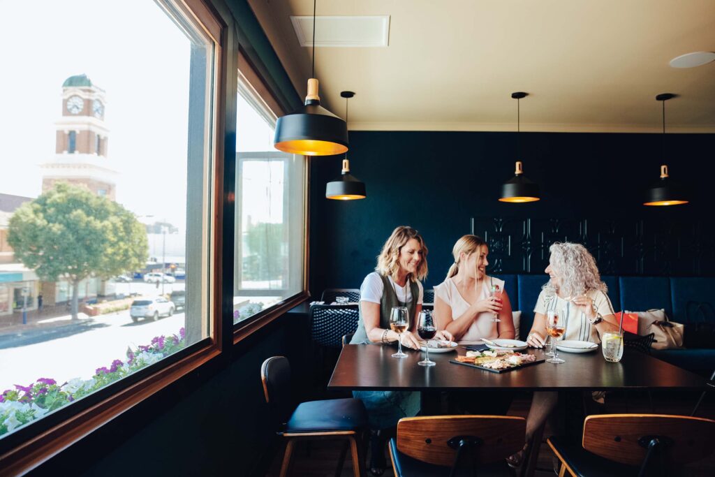 Three women sit at a table in a modern wine bar, sharing drinks and food by a large window overlooking a street with a clock tower.