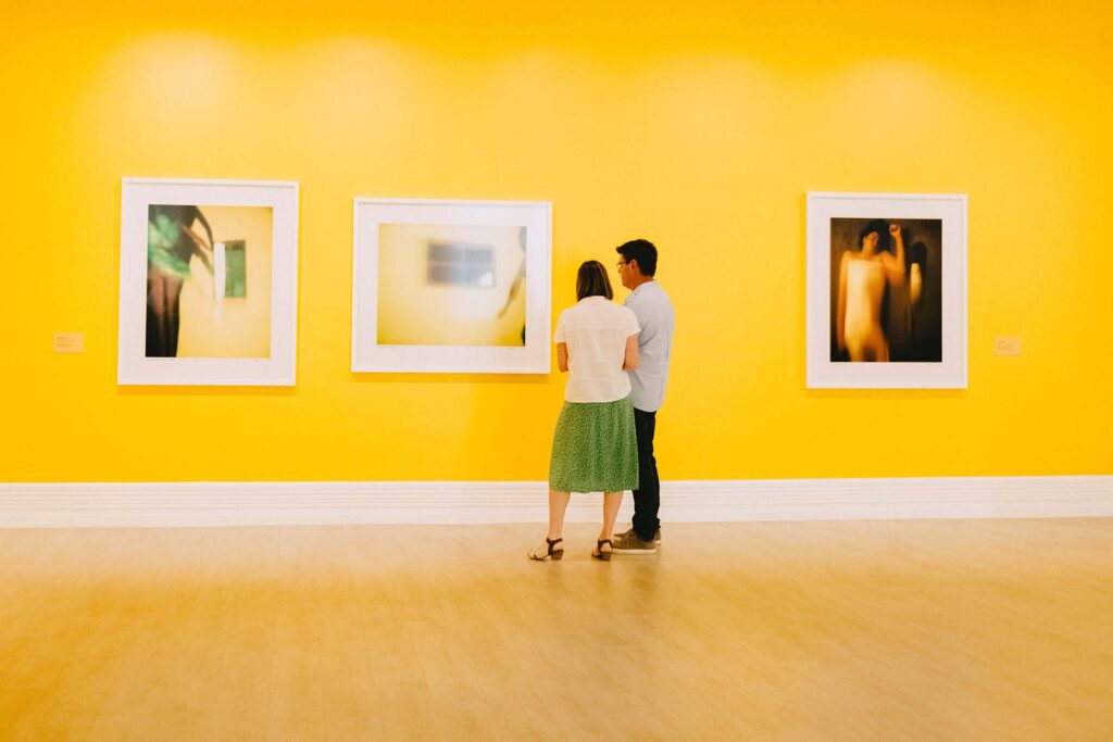 A woman in a green skirt and a man in a blue shirt with black pants look at a painting in an art gallery with bright yellow walls