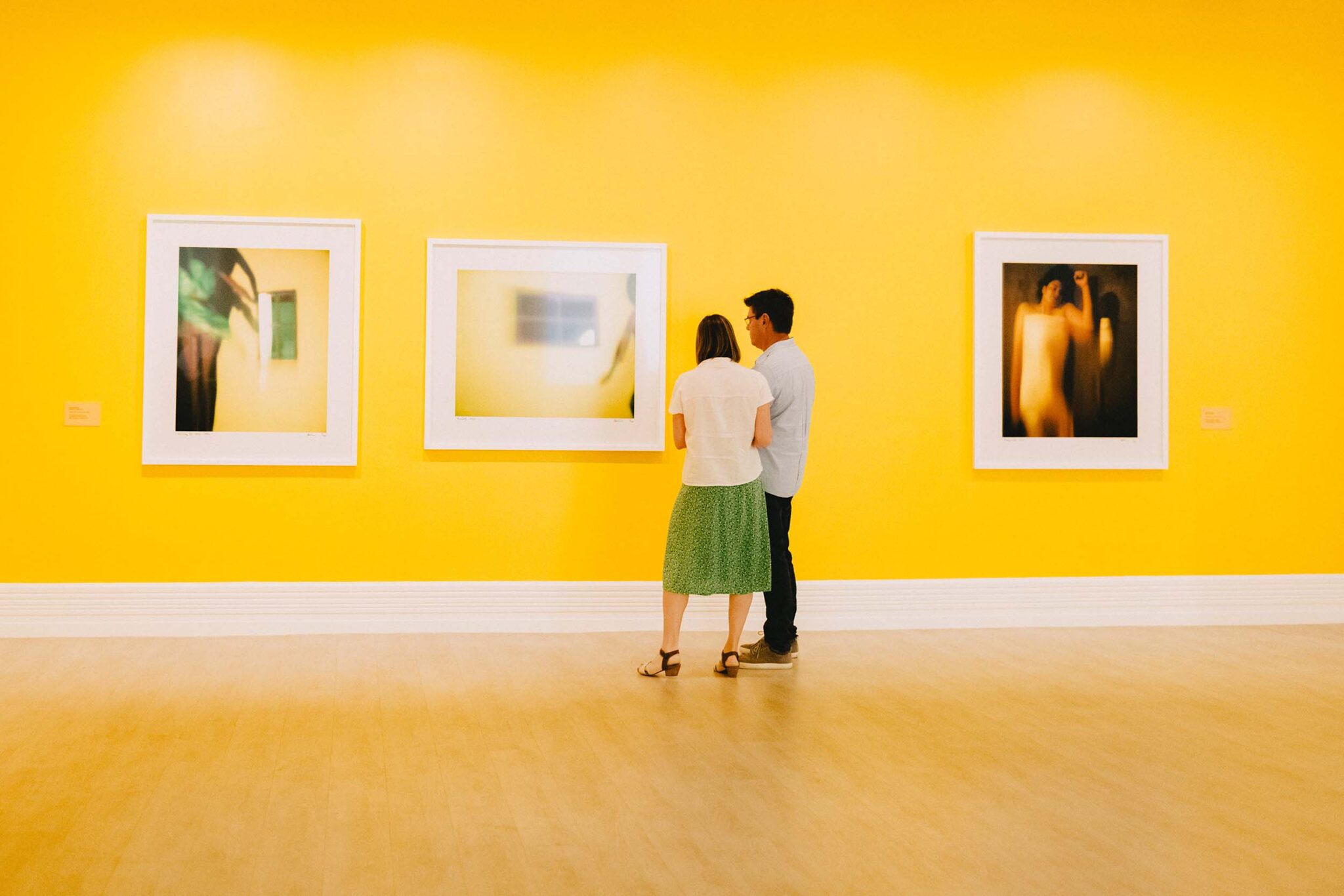 A woman in a green skirt and a man in a blue shirt with black pants look at a painting in an art gallery with bright yellow walls