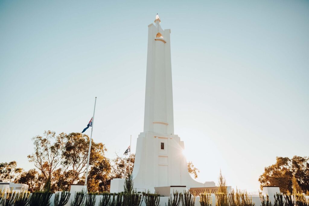 The white cenotaph of Albury's war monument is set against a pale blue sky with the sun low behind it. Australian flags fly at half-mast on nearby flag poles