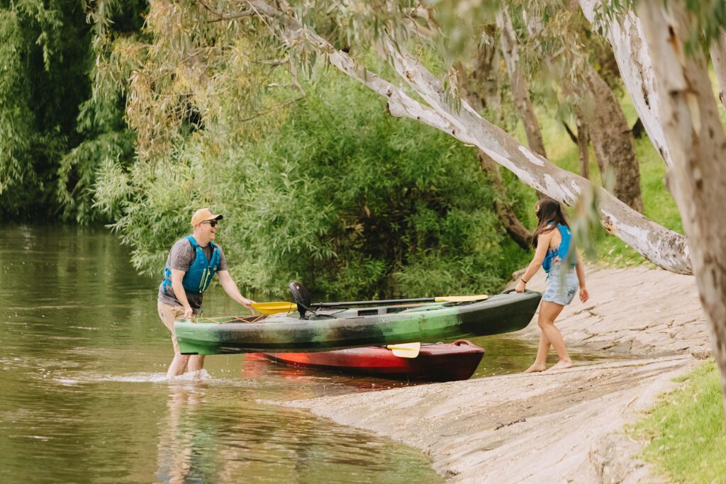 A man and woman launching a kayak into the Murray River in Albury Wodonga, surrounded by trees and calm, green water.