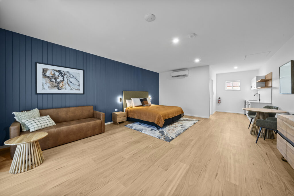 Motel room with a dark blue accent wall, king size bed, couch, table, and kitchenette with plenty of space for accessibility.