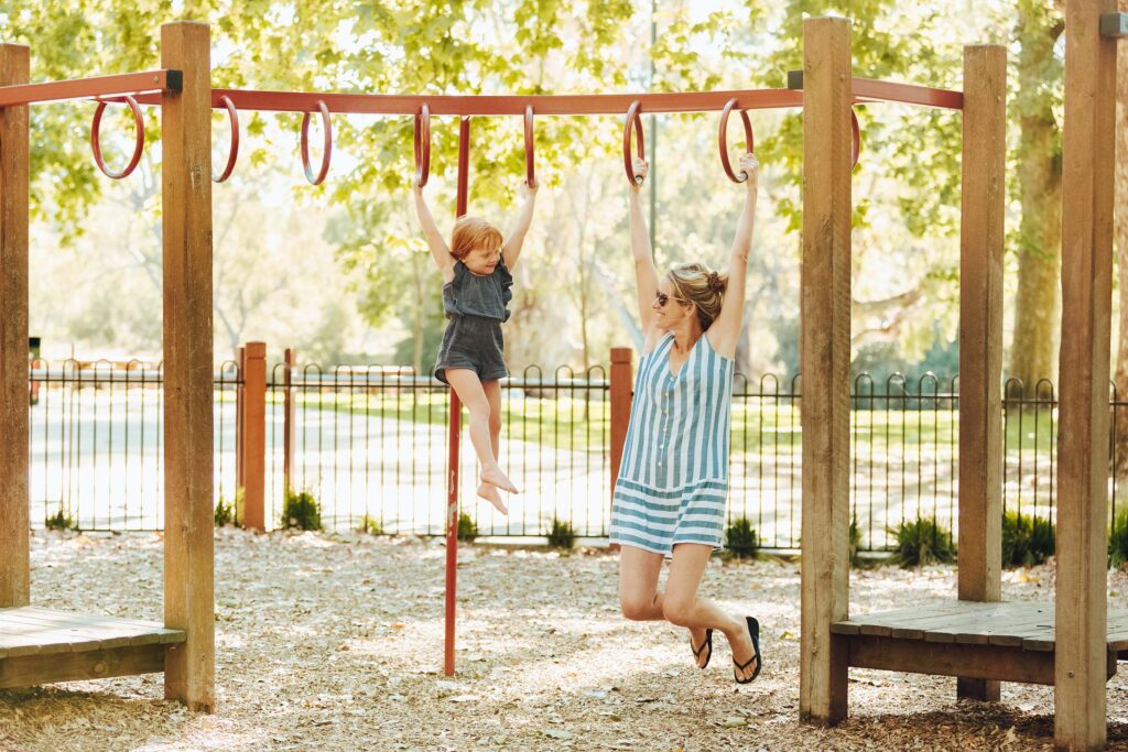 A small girl with red hair and her mother hang from the rings of a set of monkey bars inside a fenced off playground at a leafy park
