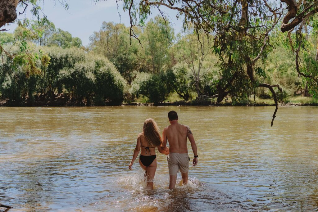 A young couple hold hands as the enter the water of the Murray River. The woman is in a black bikini with blond hair hanging down her back. The man wears beige board shorts