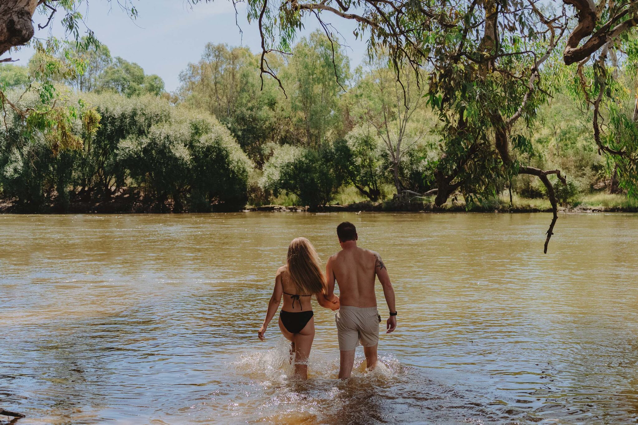 A young couple hold hands as the enter the water of the Murray River. The woman is in a black bikini with blond hair hanging down her back. The man wears beige board shorts