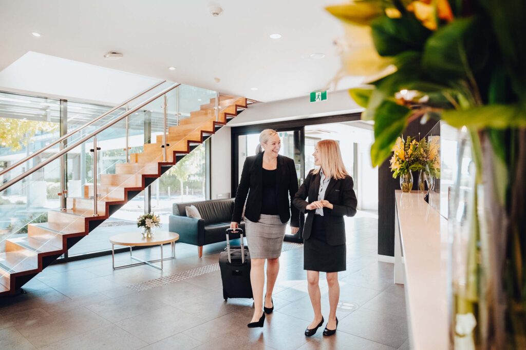 A woman in a black blazer and grey pencil skirt wheeling a suitcase is welcomed in a hotel lobby by a concierge in black blazer and skirt