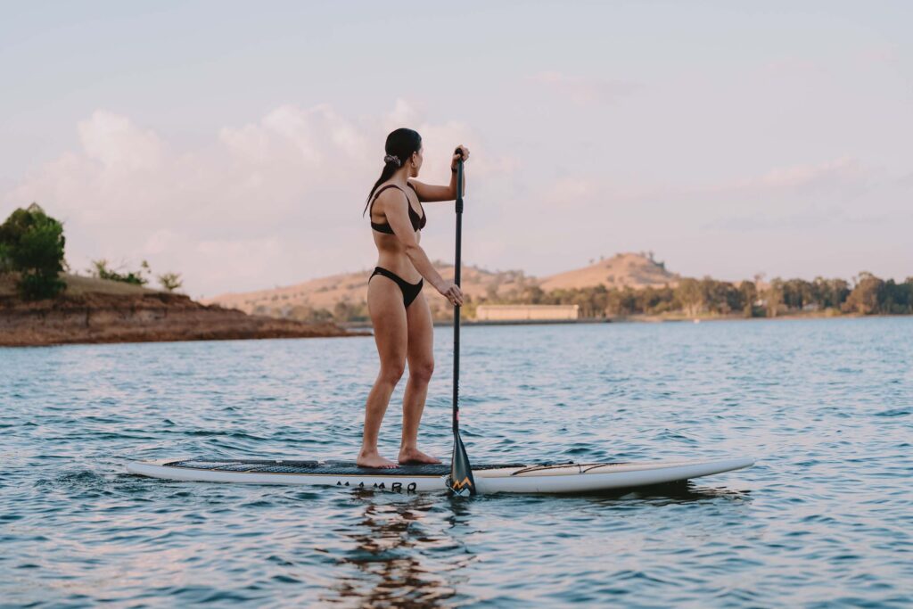 Woman in a black bikini paddleboarding on a calm lake, surrounded by gentle hills and a partly cloudy sky in the late afternoon. In the distance the Lake Hume Damn Wall can be spotted.