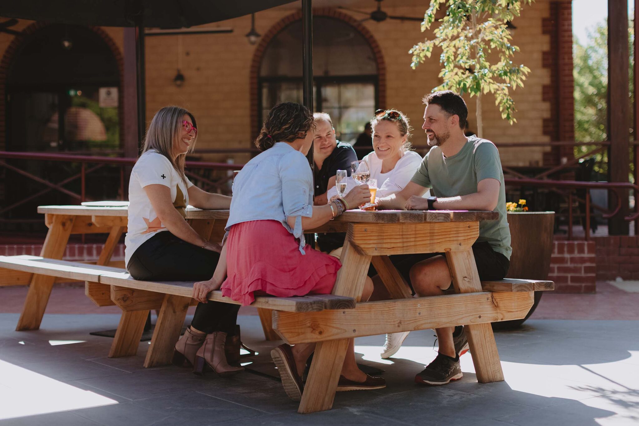 A group of friends sit at a large picnic-style table under a shade umbrella with drinks in hand. The building behind them is made of sandy-yellow bricks with red brick details, and has a large window. Sun illuminates the leaves of a small tree.