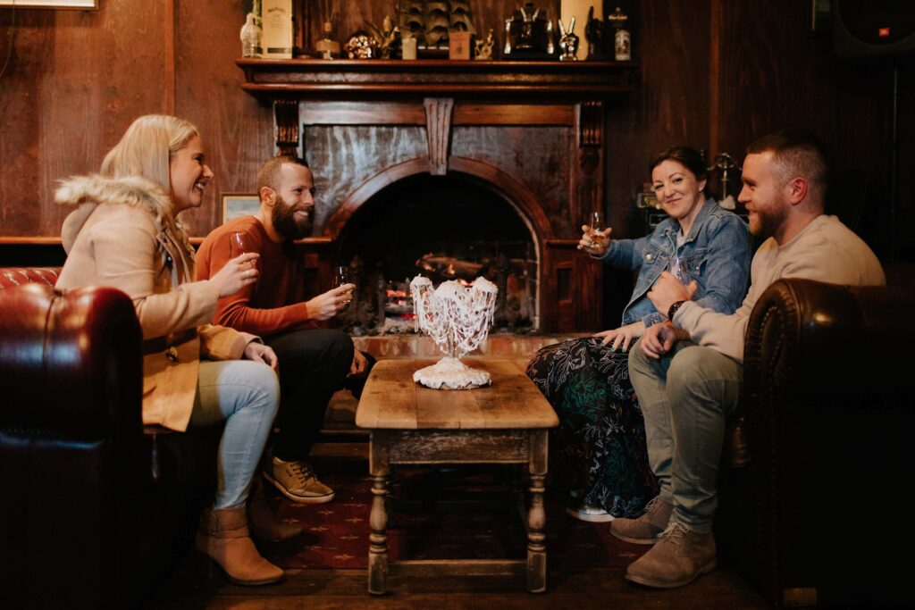 Four friends sit on leather couches around a wooden coffee table beside a cosy fireplace enjoying glasses of whisky.