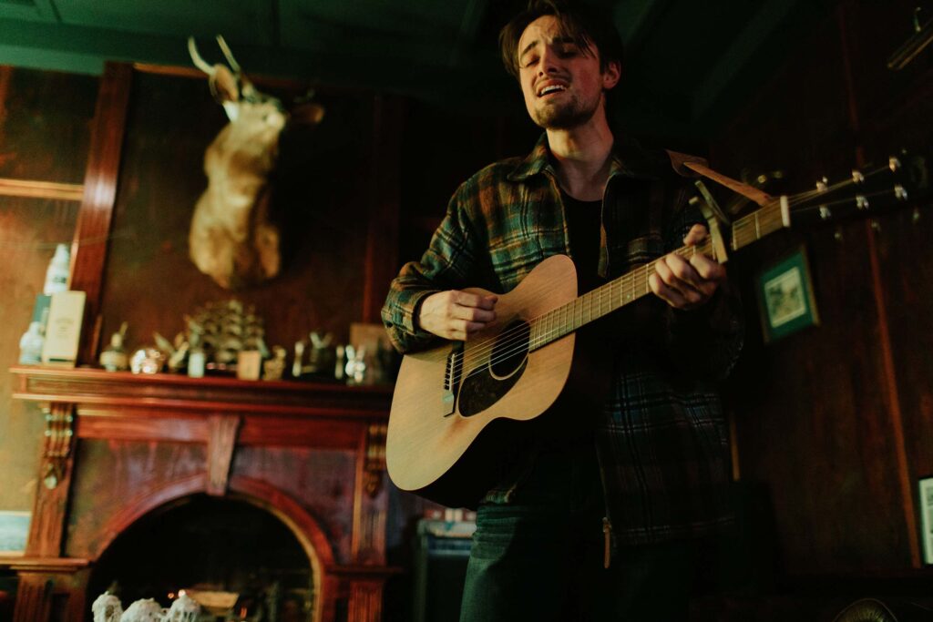 A man passionately playing an acoustic guitar indoors, in a warmly lit bar with dark wooden walls and a mounted deer head above a fireplace in the background.
