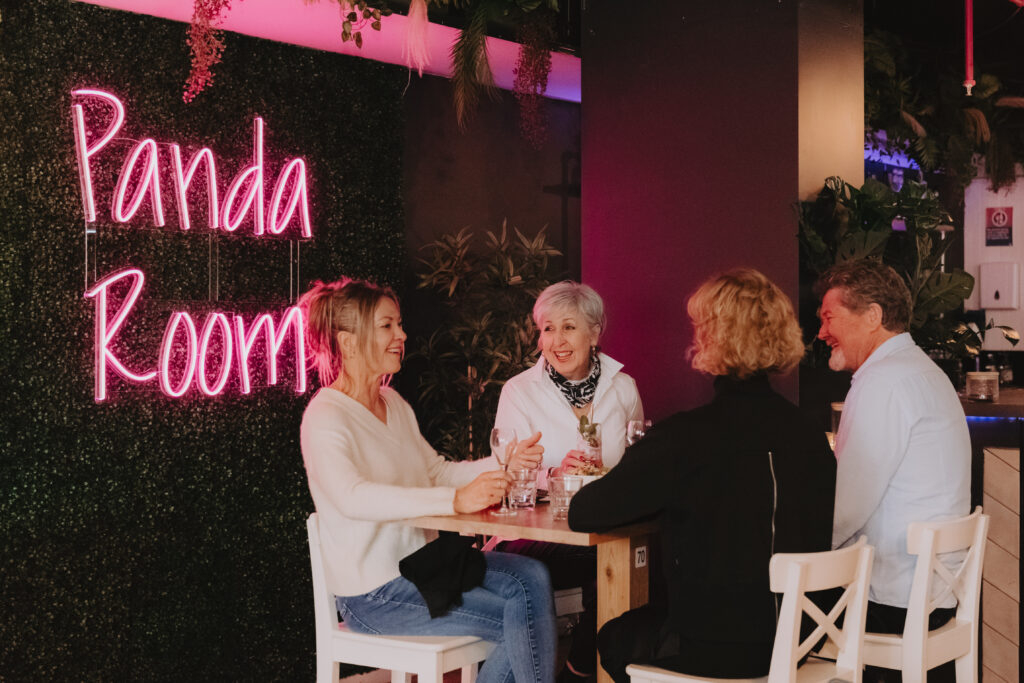 Four friends sit around a table at a restaurant. The wall is lined with faux greenery and decorated with a pink neon sign reading 'Panda Room'