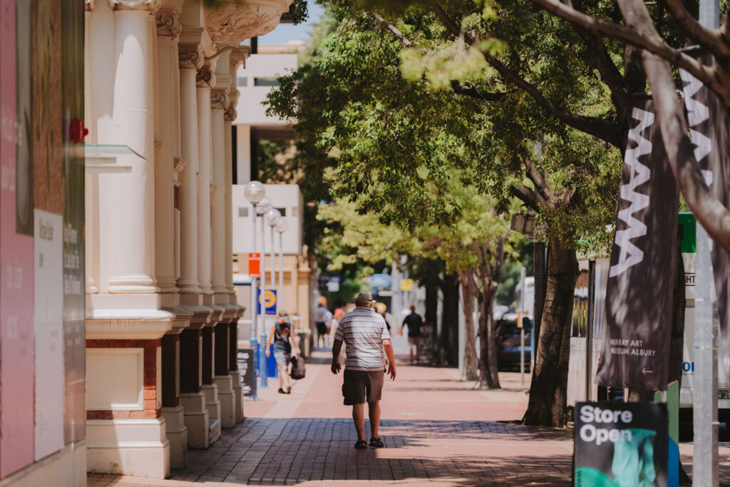 A wide brick footpath with historic buildings on the left and leafy green trees on the right. In the foreground is a flag for MAMA art gallery and a sign that reads 'store open'