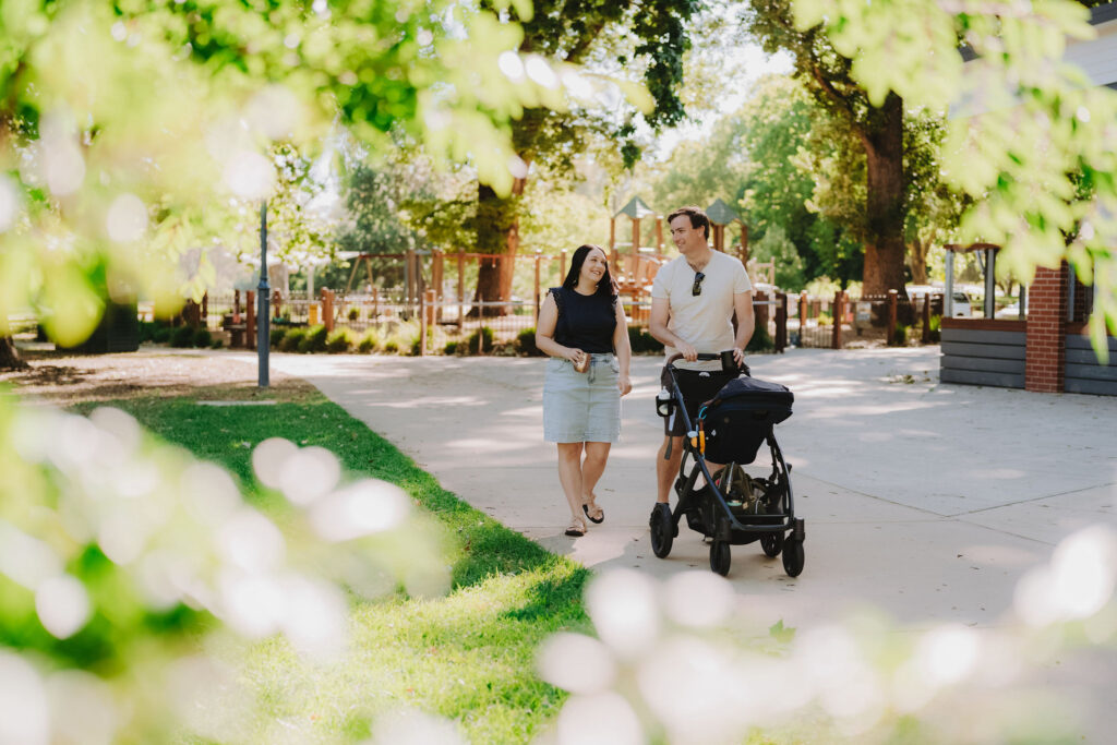 A man in a white t-shirt pushes a pram in the morning sun alongside a woman in a navy blouse and denim skirt. There is a playground in the background and green grass beside the path. The image is framed by sunlit foliage.